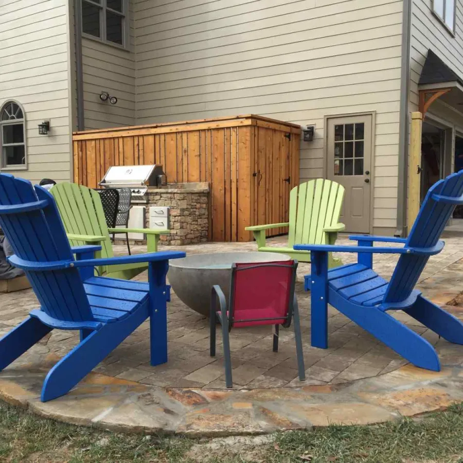 Outdoor patio with blue and green Adirondack chairs around a fire pit near a wooden house and grill setup.