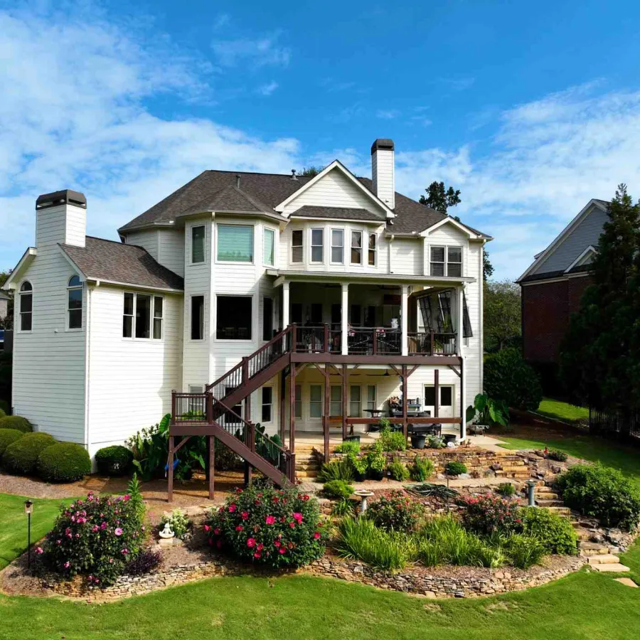 Large white two-story house with wooden deck, outdoor stairs, and landscaped garden under blue sky.