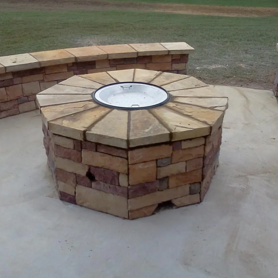 Stone outdoor fire pit with curved stone seating on concrete patio in a grassy backyard.