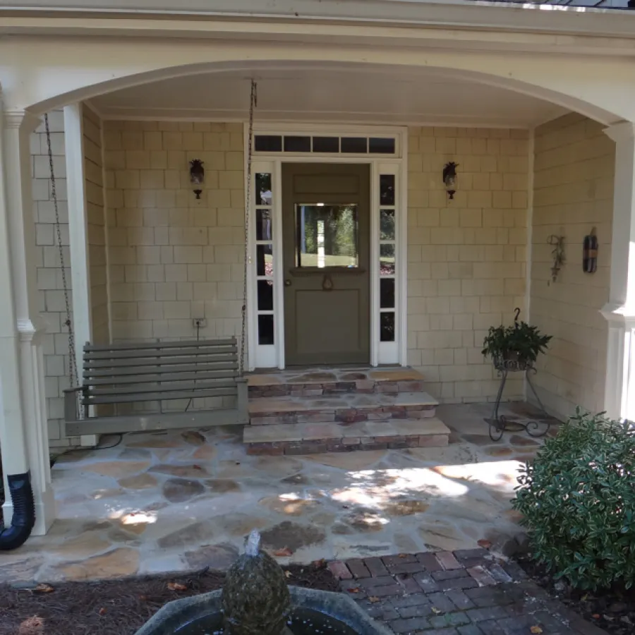 Covered porch with stone floor, wooden swing, green door, and potted plant near entrance steps