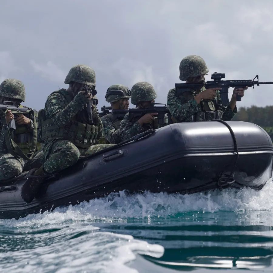 Soldiers in camouflage gear aiming rifles while riding a black inflatable boat on water during training.