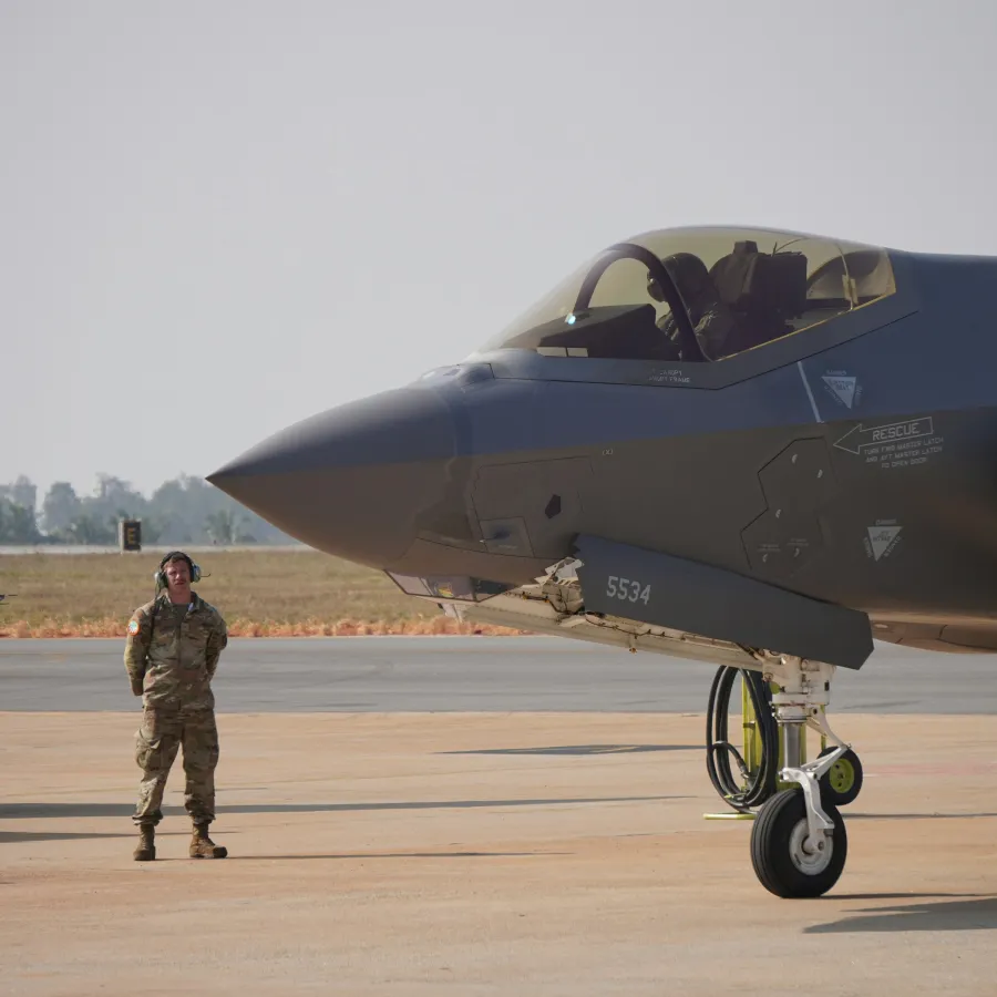 Military personnel standing next to a parked stealth fighter jet on an airfield during daytime