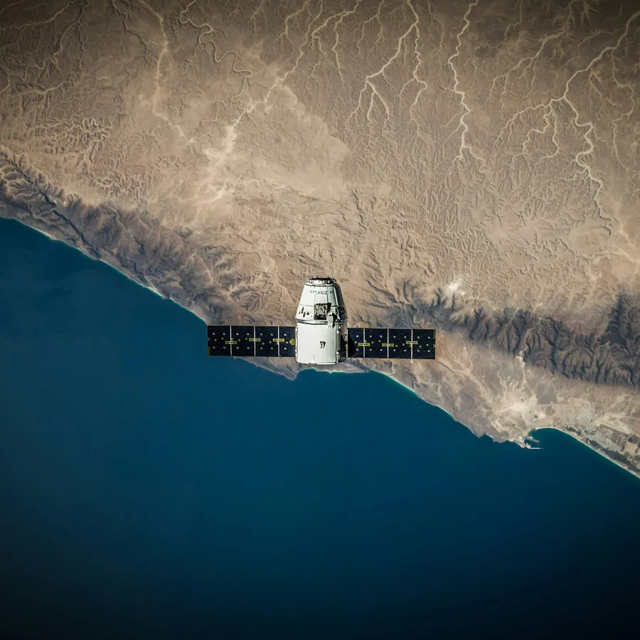 Satellite with solar panels orbiting over coastal desert and deep blue ocean beneath clear sky.