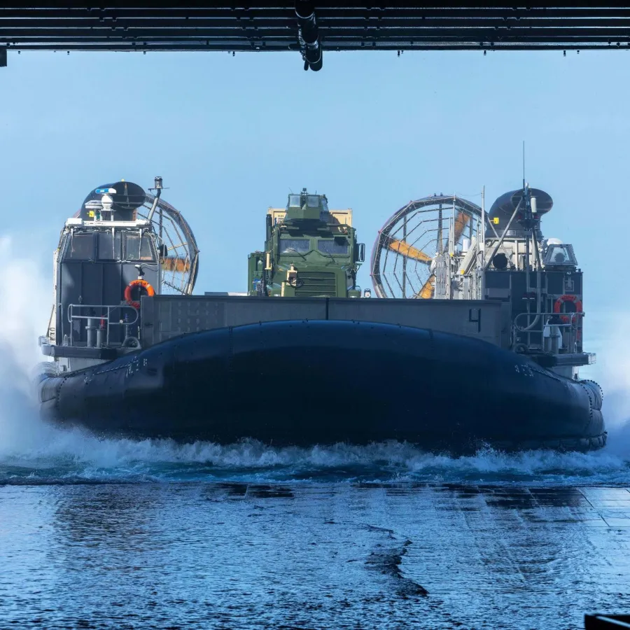 Military hovercraft launching from ship ramp creating water spray under blue sky.
