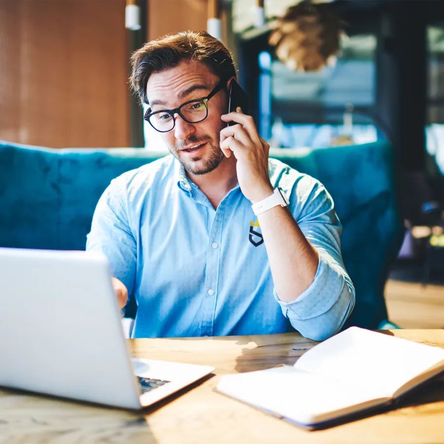Man wearing glasses talks on phone while working on laptop at wooden desk with notebook open