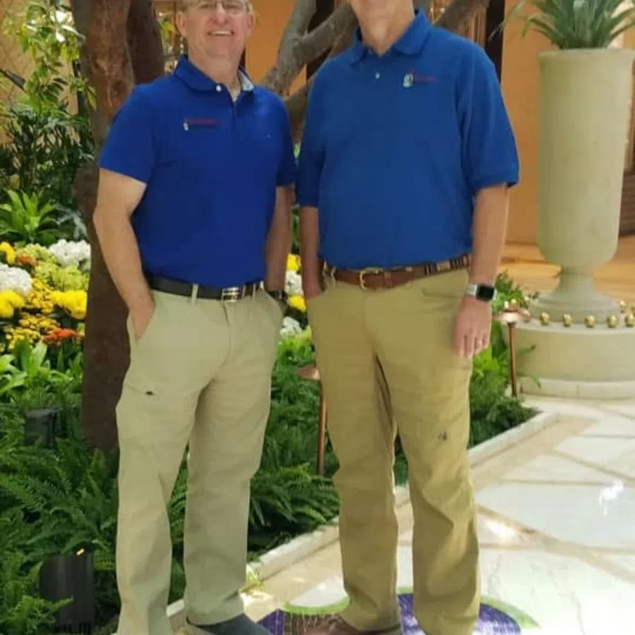 Two men in blue polo shirts and khaki pants standing indoors with plants and decorative flooring.