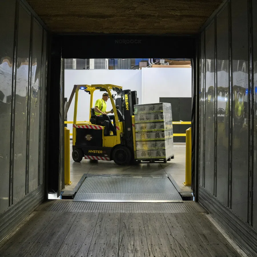 Forklift operator loading stacked pallets into a trailer through a warehouse loading dock with metal walls.