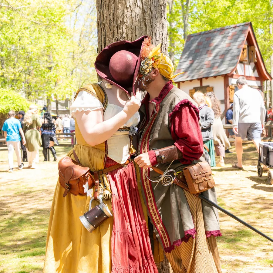 Couple in colorful Renaissance costumes sharing a secret behind a hat at a sunny outdoor festival.