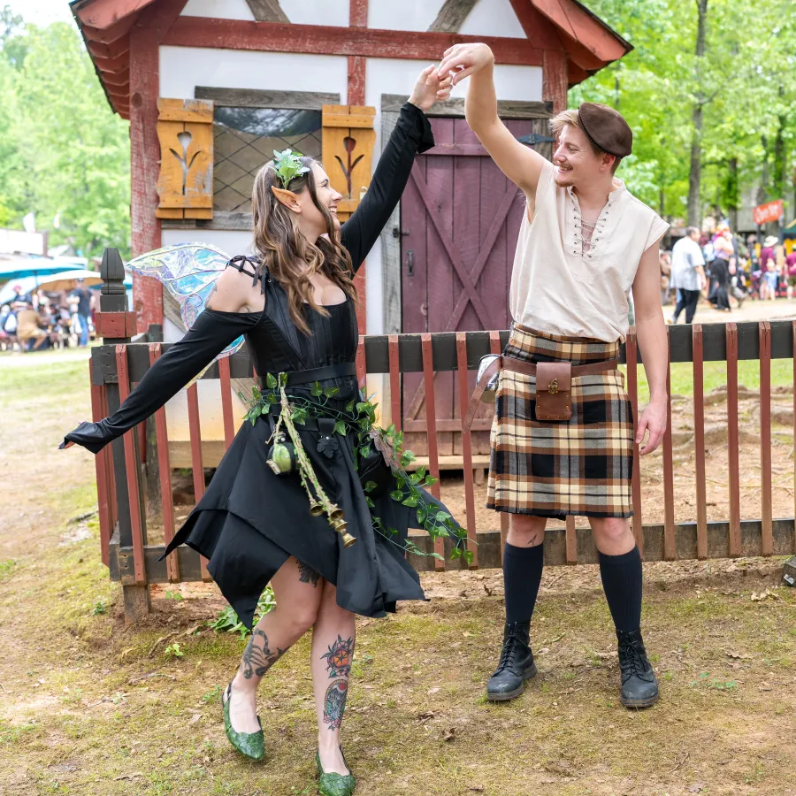 Man in kilt and woman dressed as fairy dancing outdoors at a Renaissance fair with rustic wooden building background