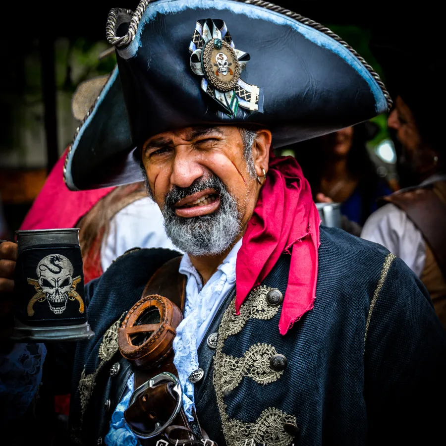 Middle-aged man dressed as a pirate with a black tricorn hat, red scarf, ornate coat, holding a skull-decorated mug.