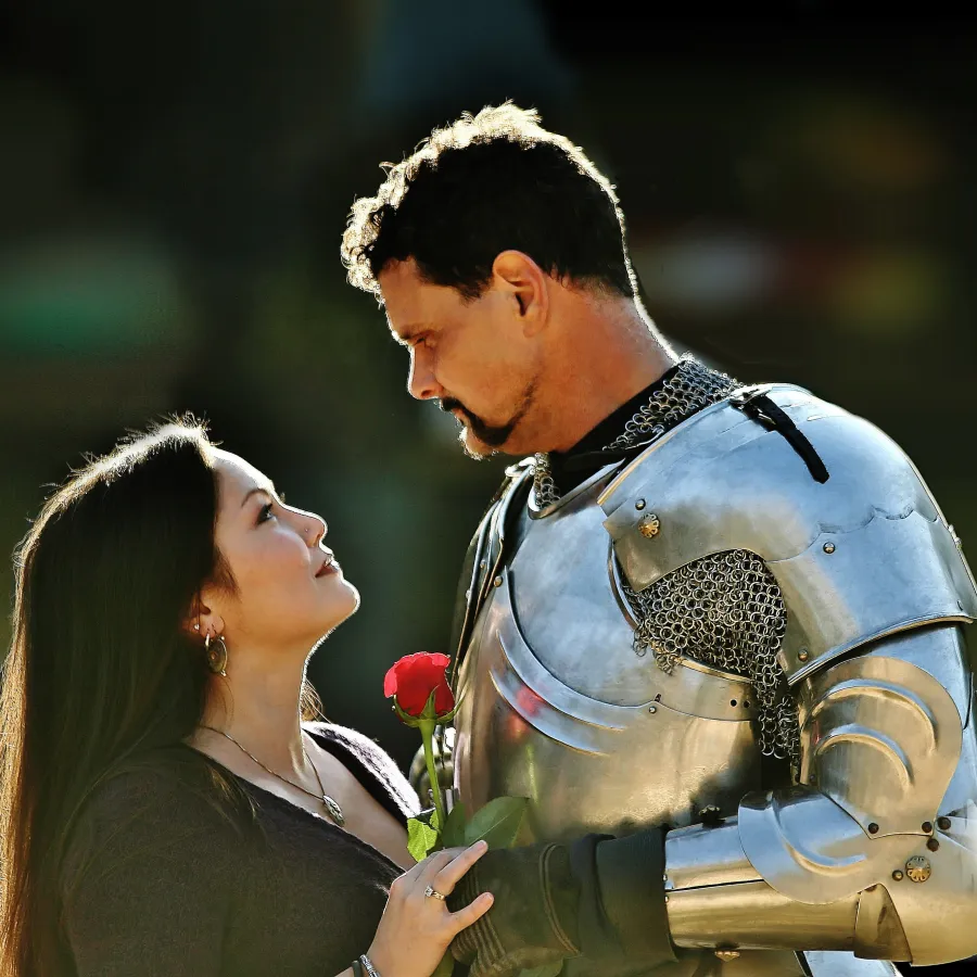 Woman gazes up at man in shining medieval armor holding a red rose, both sharing an intimate moment.