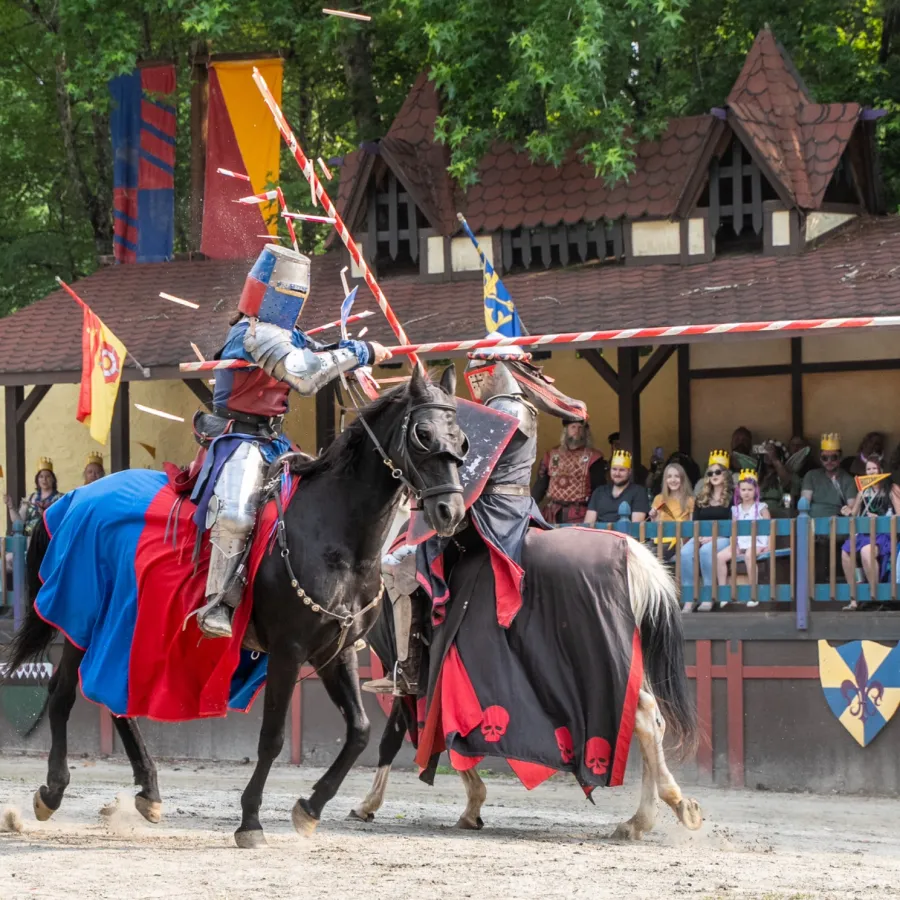 Two knights in armor jousting on horses with spectators watching at a medieval festival event.