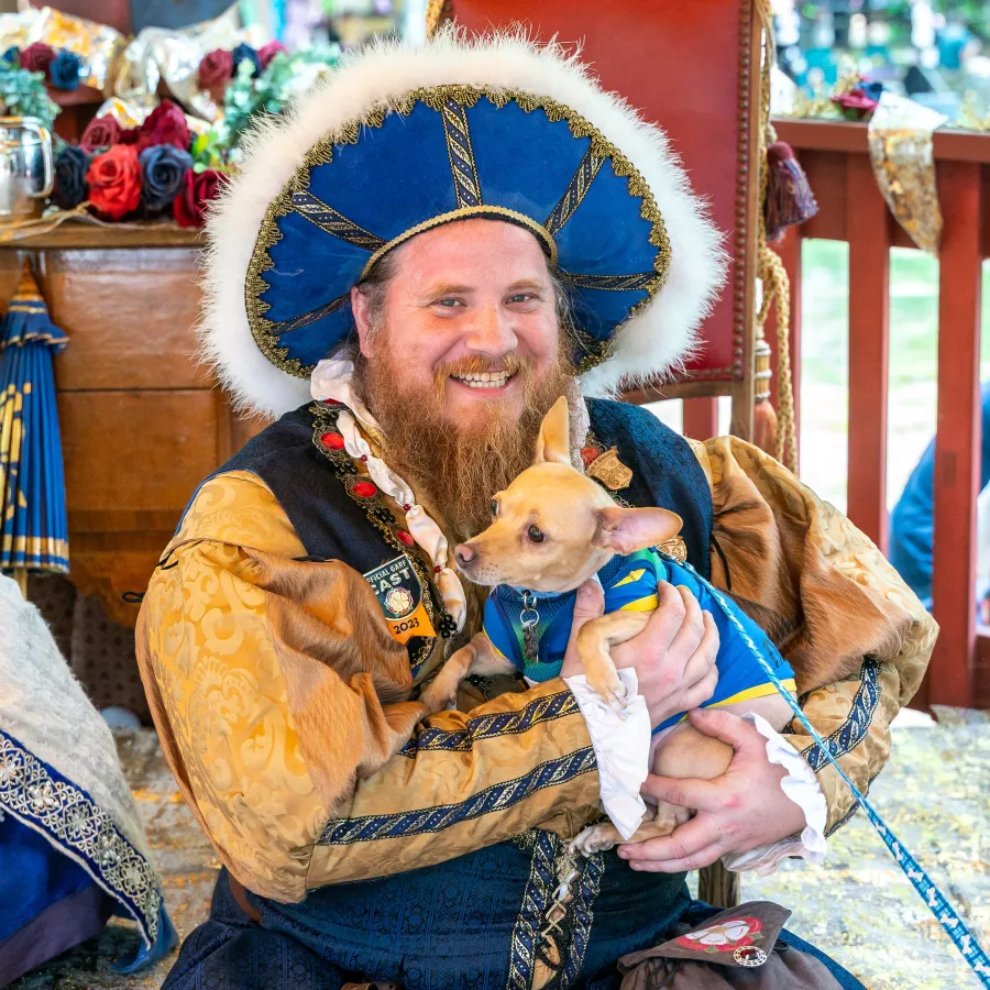 Smiling man in Renaissance costume holds small dog wearing blue outfit at outdoor event.