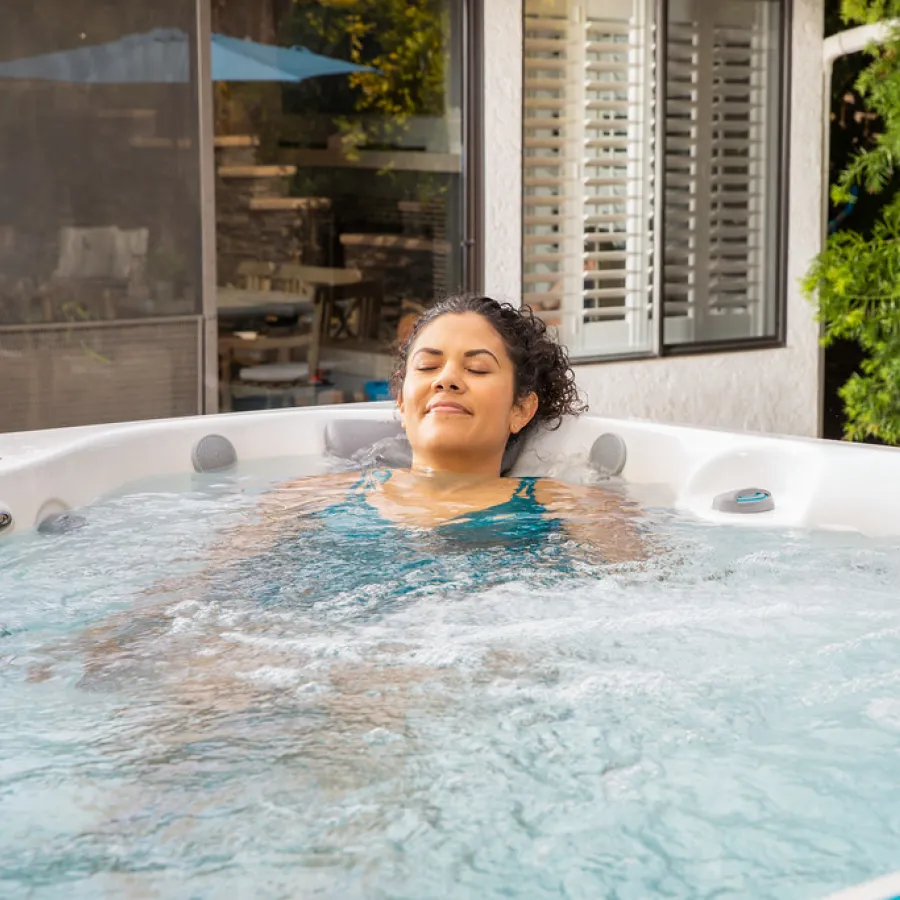 Woman relaxing in a bubbling outdoor hot tub near a house with greenery and patio furniture.