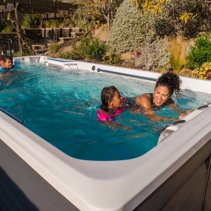 Family enjoying swimming and playing in a large outdoor swim spa surrounded by greenery.
