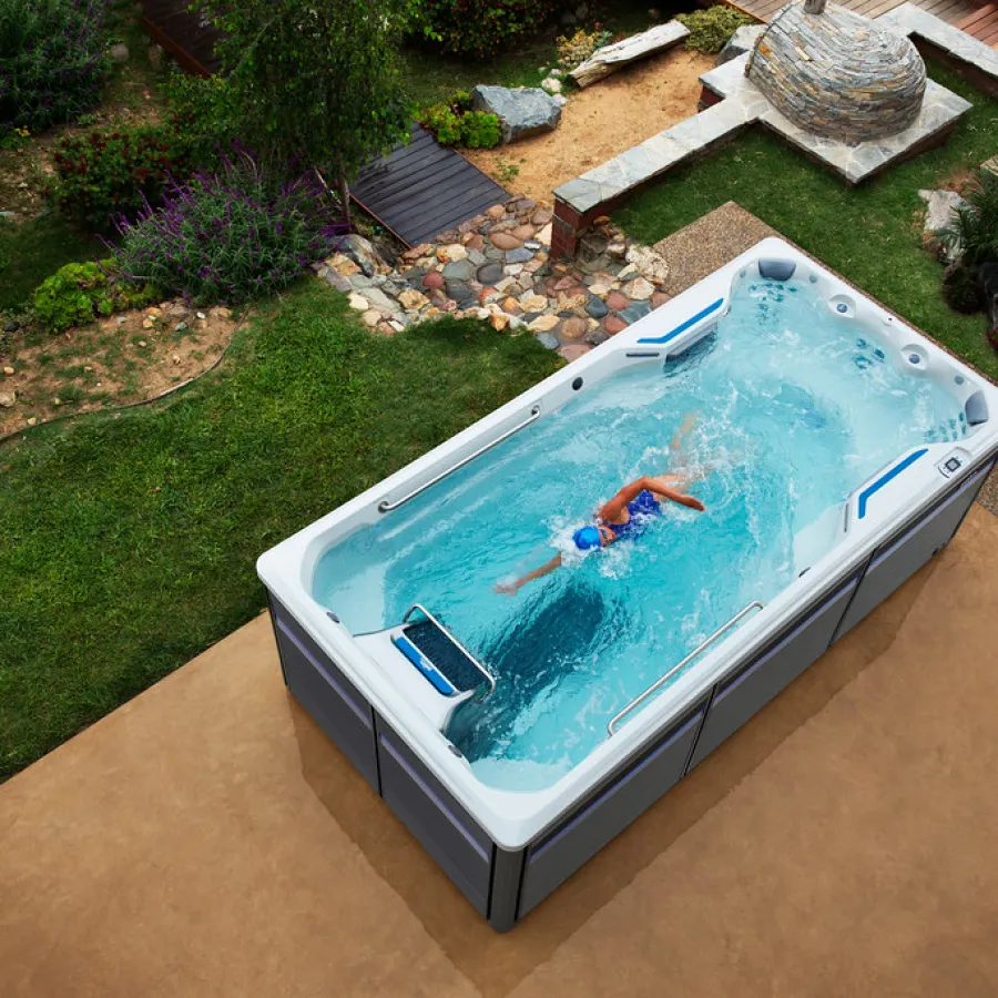 Swimmer exercising in a modern swim spa installed in a landscaped backyard with greenery and stone paths.