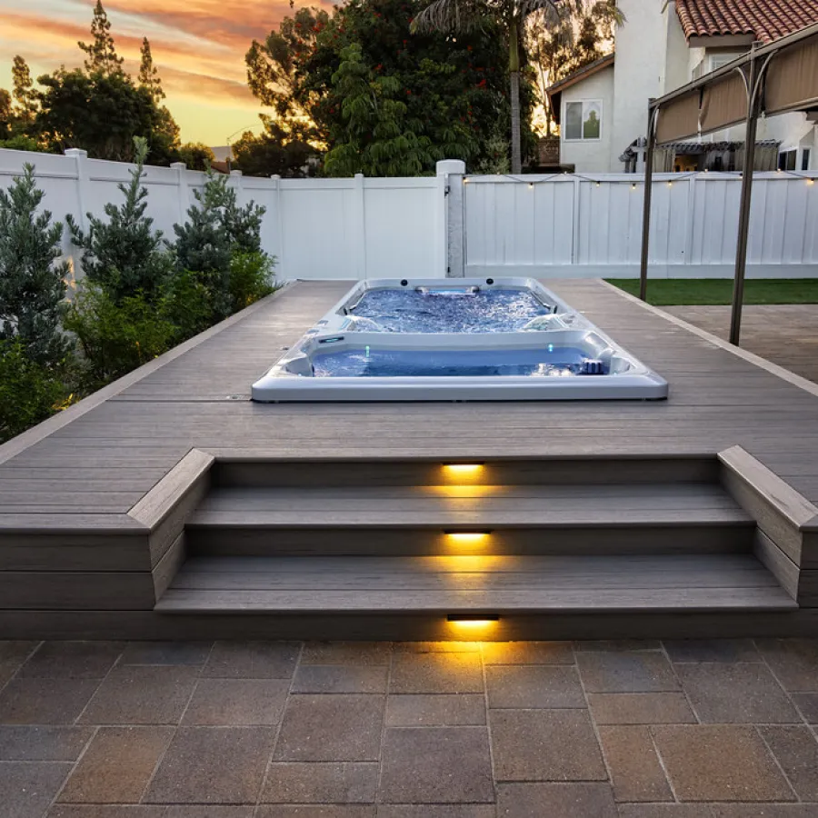 Modern backyard with an elevated swimming pool, wooden deck, ambient step lighting, and a sunset sky.