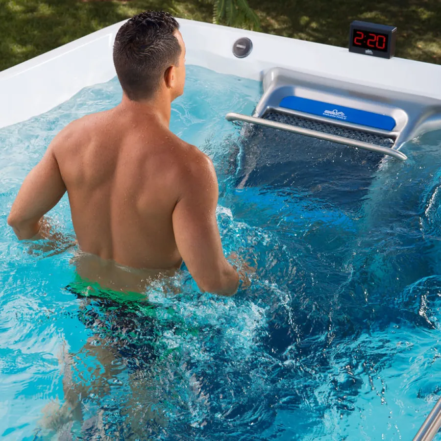 Man swimming against current in an outdoor swim spa with digital timer and clear blue water.