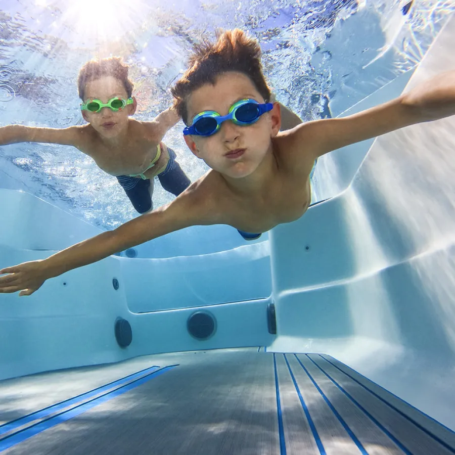 Two boys wearing goggles swimming underwater in a clear pool with light reflections