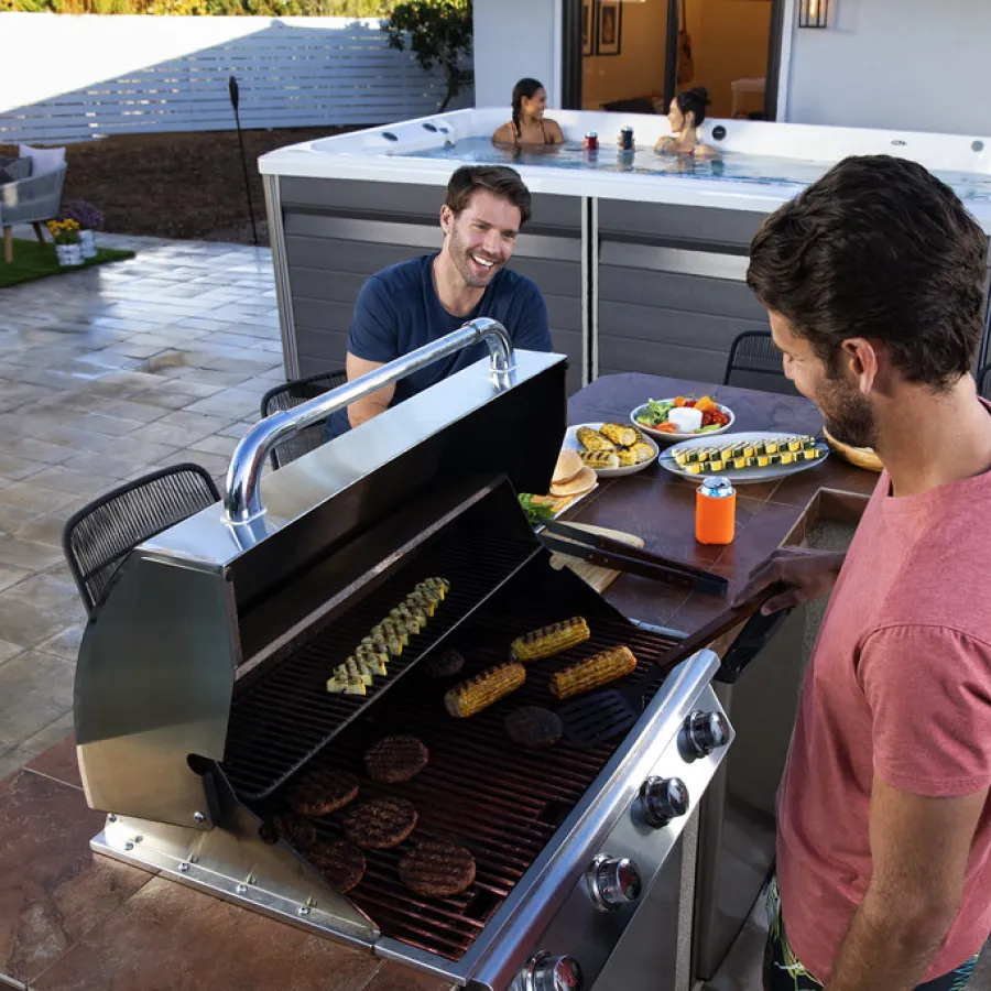 Two men grilling food on an outdoor barbecue near a hot tub with two people relaxing inside at sunset.