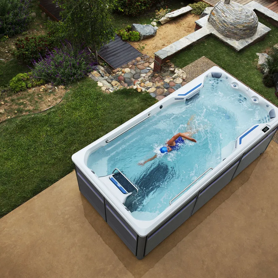 A swimmer doing laps in a modern rectangular swim spa set in a landscaped backyard with greenery and stone pathways.