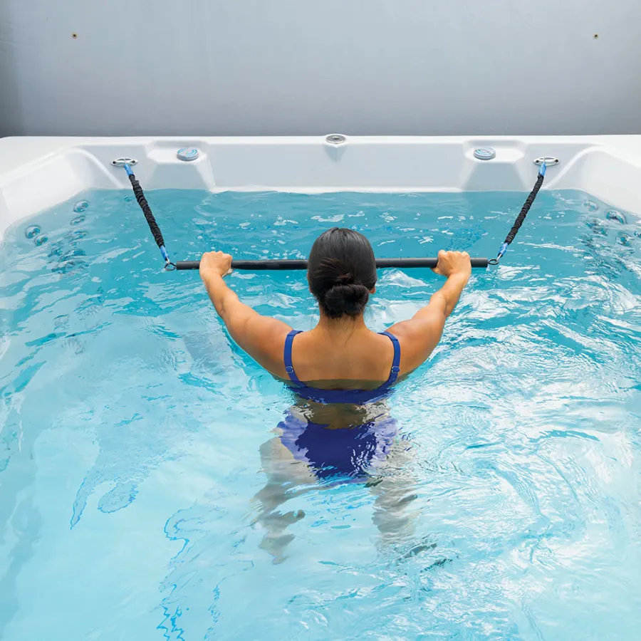Woman in blue swimsuit exercising with resistance bands in a clear water pool from a rear view.