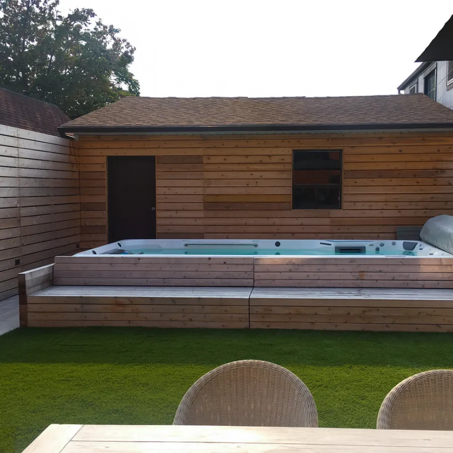 Modern backyard with wooden hot tub enclosure, artificial grass, and wooden shed under clear sky.