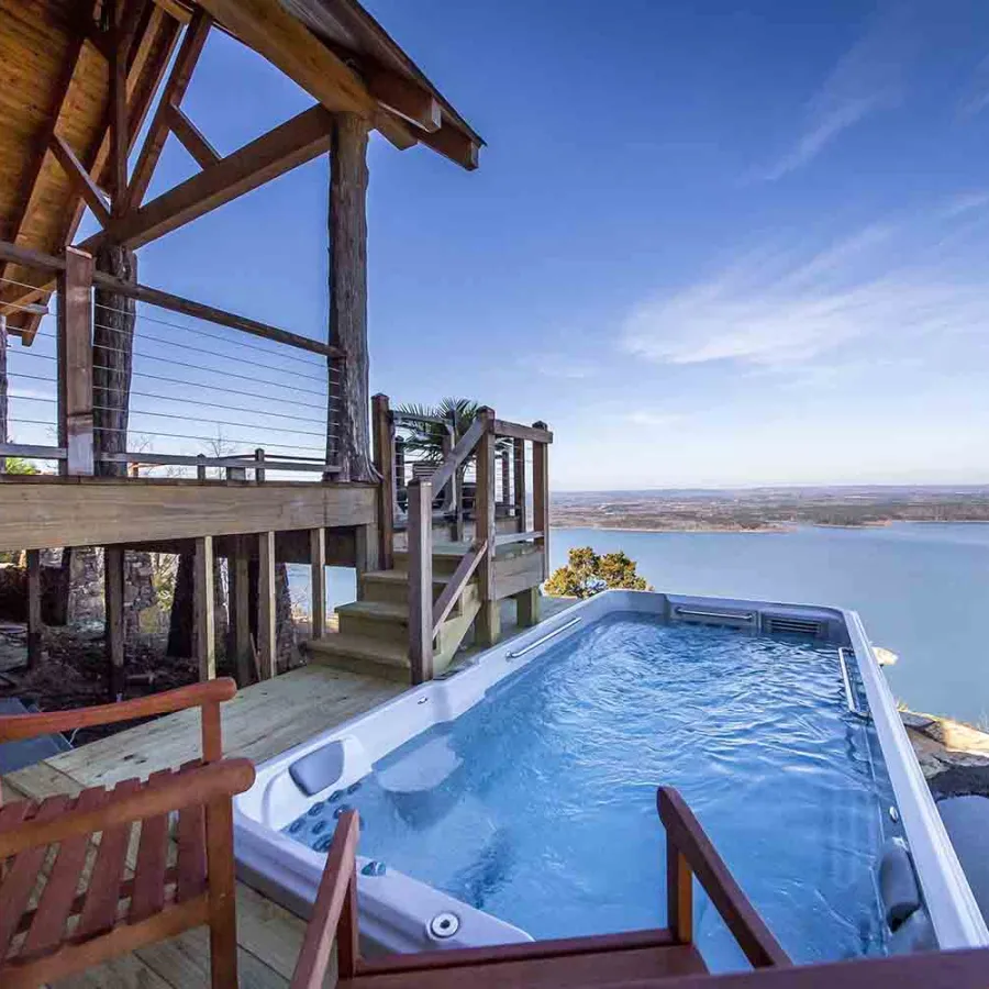 Outdoor hot tub on wooden deck overlooking lake and distant landscape under clear blue sky.