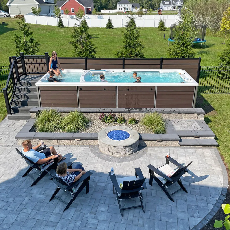 Backyard with above-ground pool, children swimming, adults relaxing on patio chairs around fire pit on sunny day.
