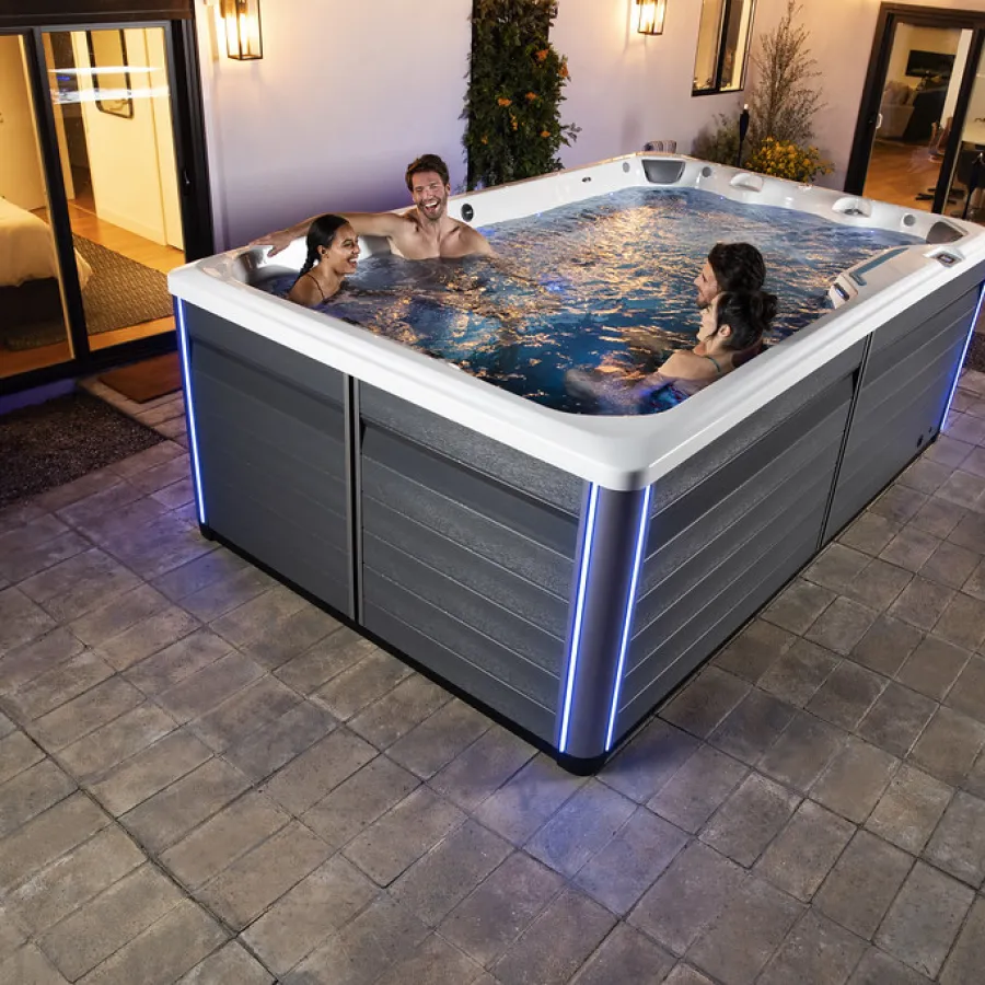 Four friends enjoying a modern outdoor hot tub with LED lights at night on a tiled patio near a house.