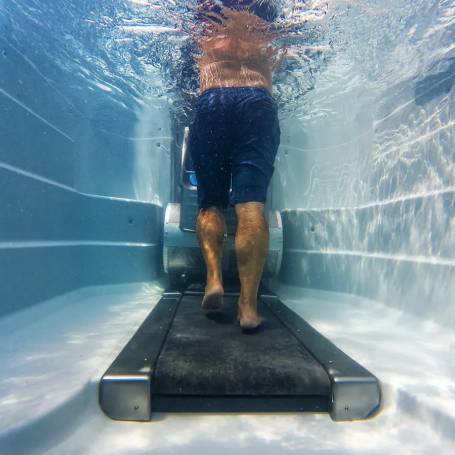 Underwater view of a man walking on a treadmill submerged in a pool with water reflections.