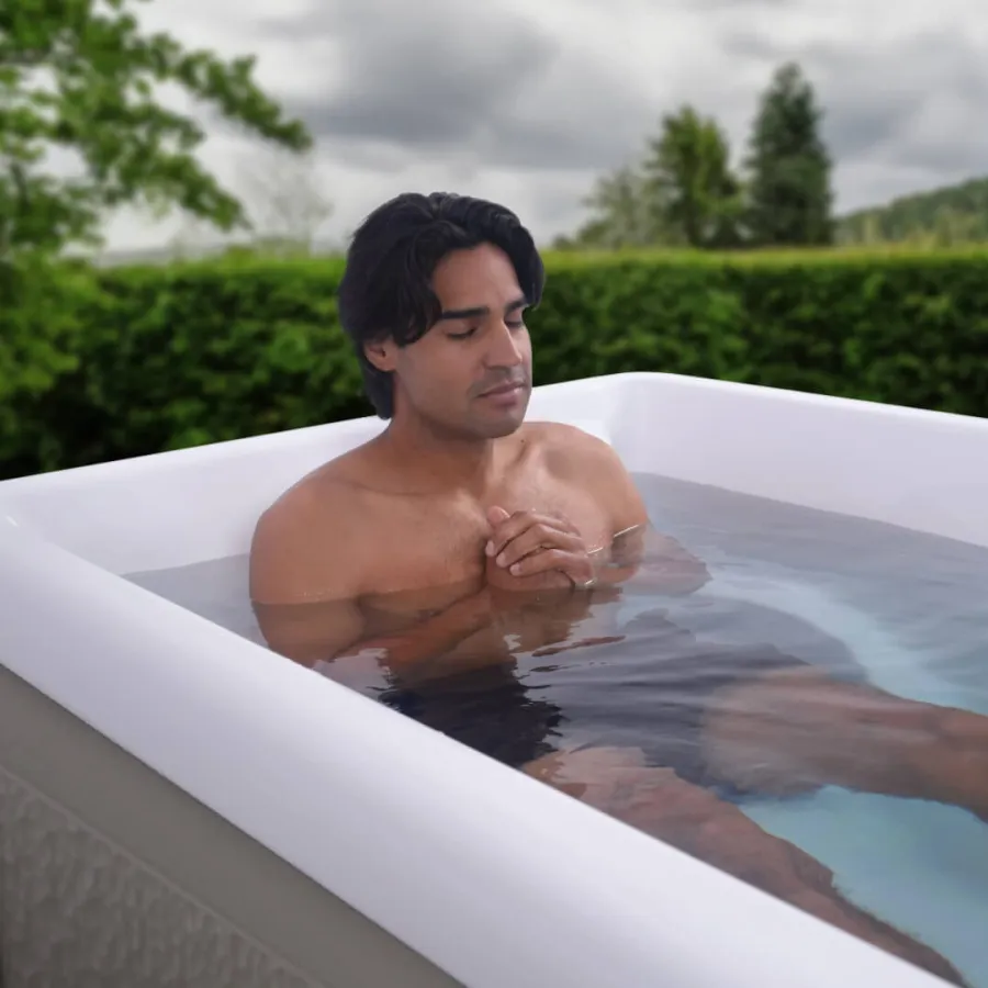 Young man relaxing with closed eyes in an outdoor hot tub surrounded by greenery and cloudy sky.
