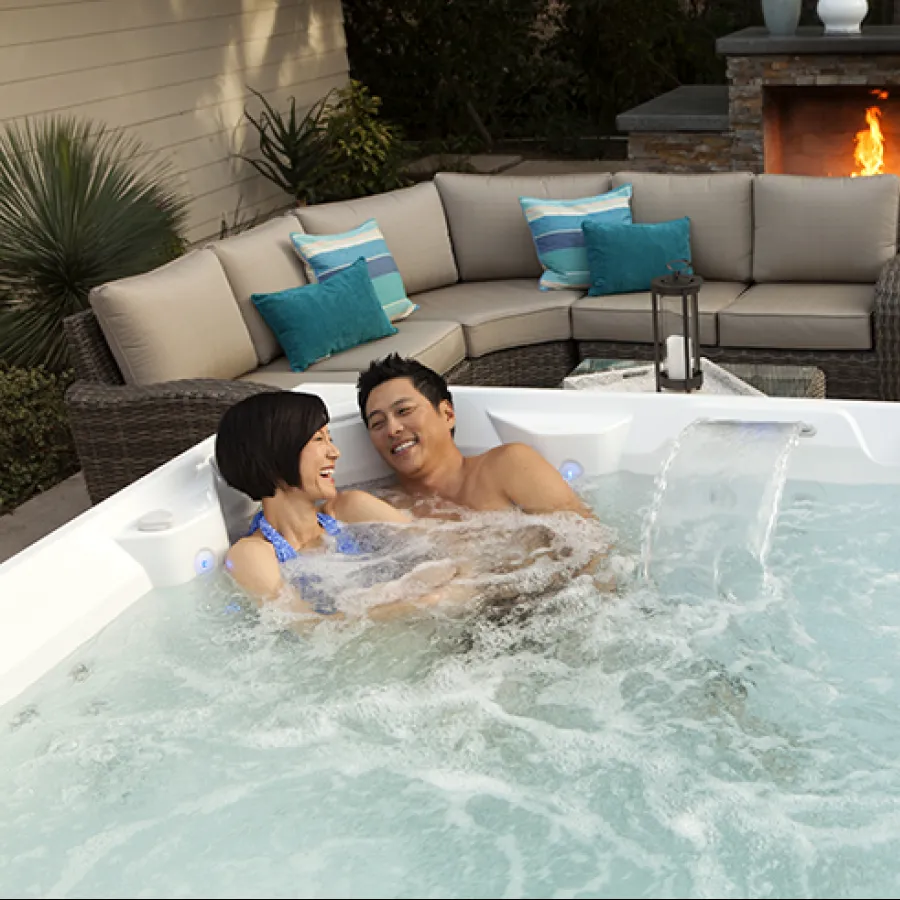 Couple enjoying a hot tub with bubbling water next to outdoor seating and a lit stone fireplace.
