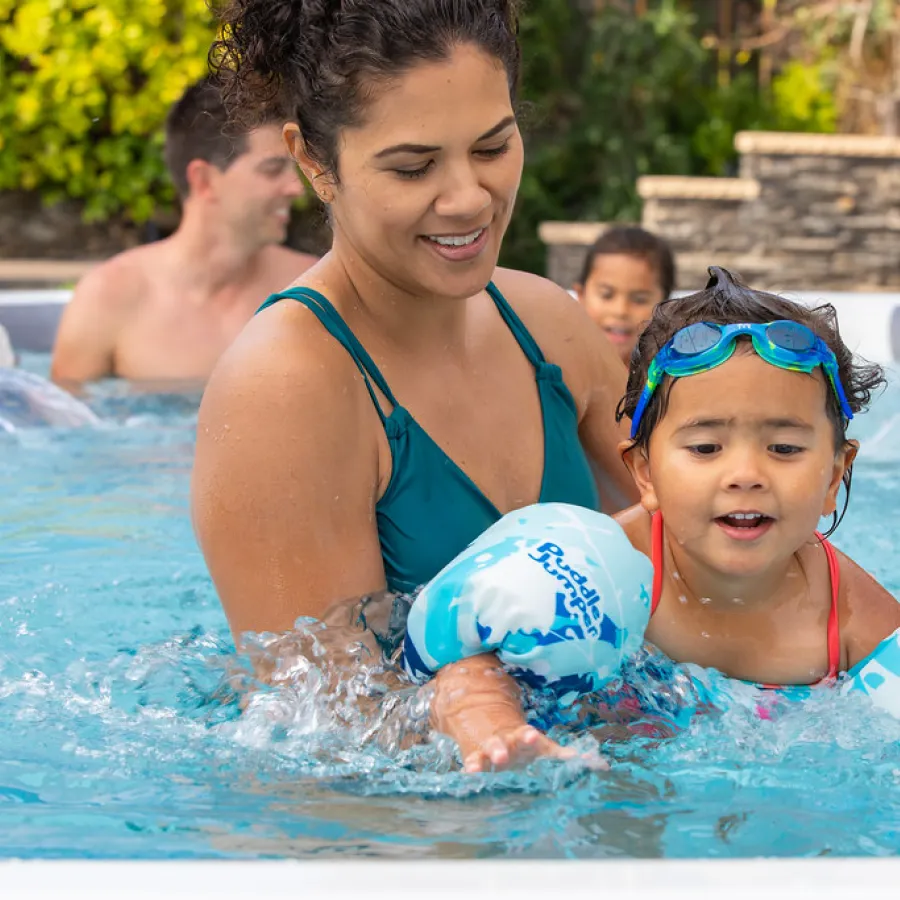 Mother helps young child with swim floaties in pool while family enjoys water activity outdoors.