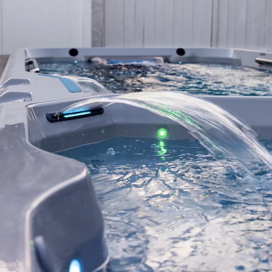 Outdoor hot tub with flowing water feature and glowing LED lights on a deck by white fence