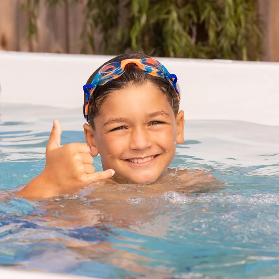 Happy boy wearing swimming goggles smiling and making shaka sign in pool water near wooden fence.