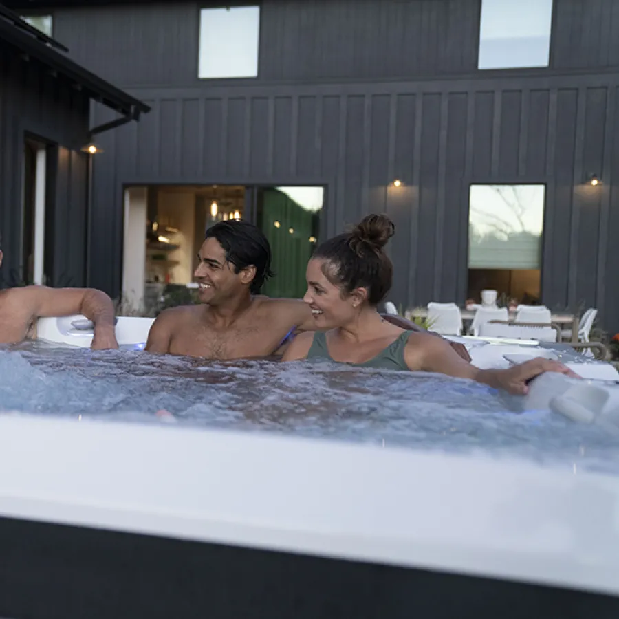 Four adults enjoying a relaxing evening in an outdoor hot tub near a modern house with dark siding.