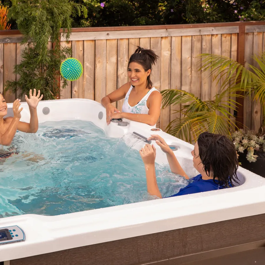 Three children playing with a ball inside a backyard hot tub surrounded by plants and wooden fence.