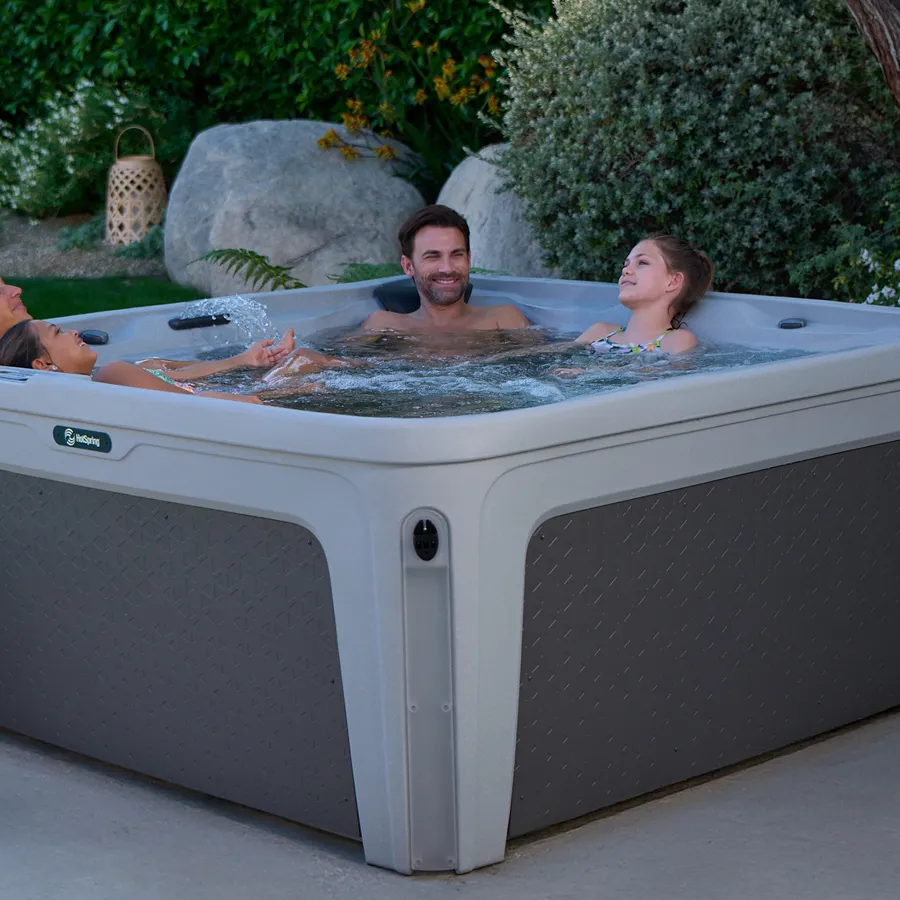Four people relaxing and enjoying a gray outdoor hot tub in a garden setting during evening.
