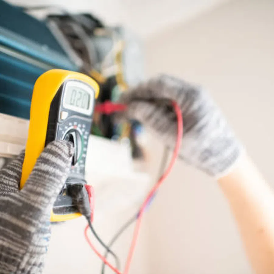 Technician wearing gloves testing an air conditioner unit with a digital multimeter for voltage readings.