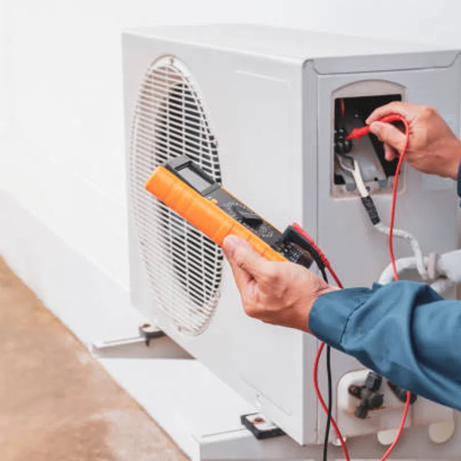 Technician using a multimeter to test an air conditioning unit outdoors with blue uniform and tools.