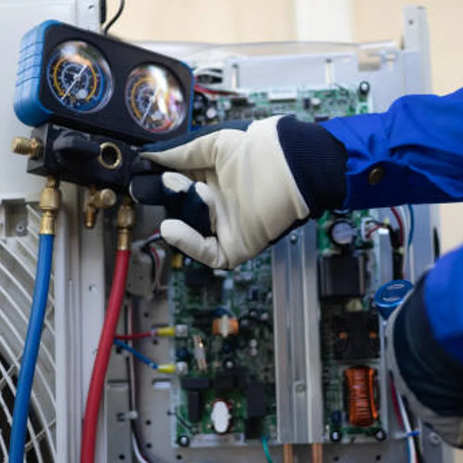 Technician adjusting gauges on an air conditioning unit with control circuit board visible, wearing protective gloves.