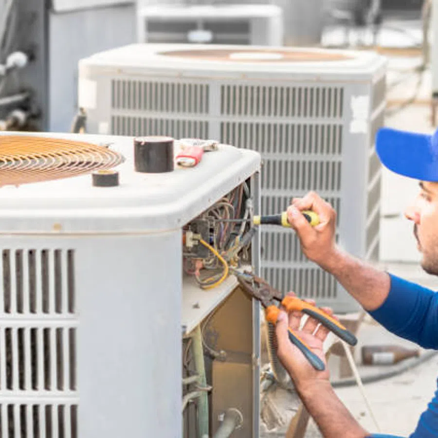 Technician in blue cap repairing air conditioning unit outdoors using screwdriver and pliers