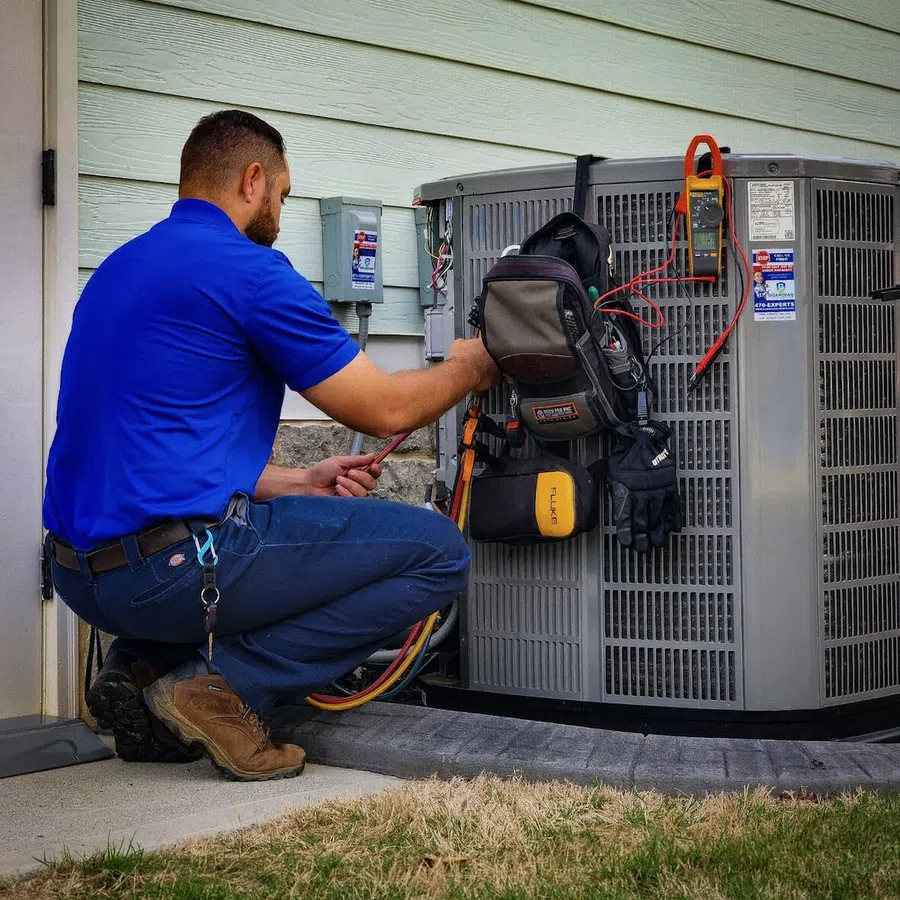 Technician in blue shirt repairing an outdoor air conditioning unit with tools and multimeter attached.