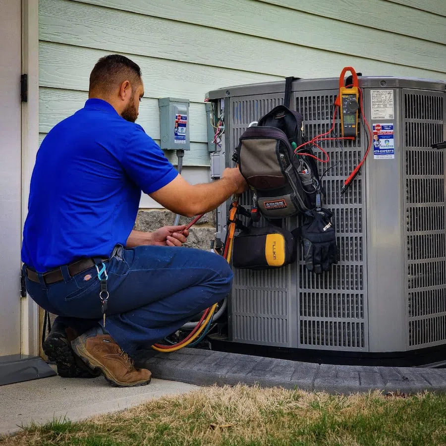 Technician in blue uniform repairing air conditioning unit outdoors with tools and equipment.