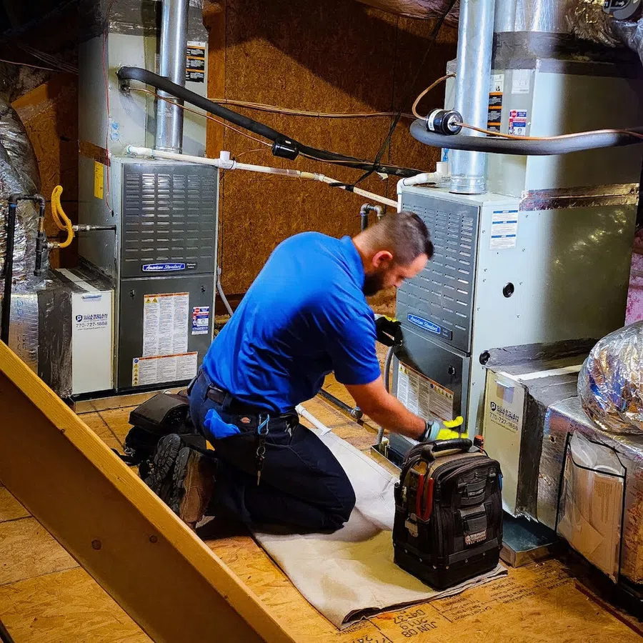 Technician in blue shirt kneeling and servicing HVAC units in a basement with tools and insulation visible.