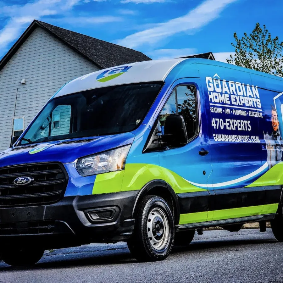 Blue and green Guardian Home Experts service van parked on a residential street in front of houses under a blue sky.