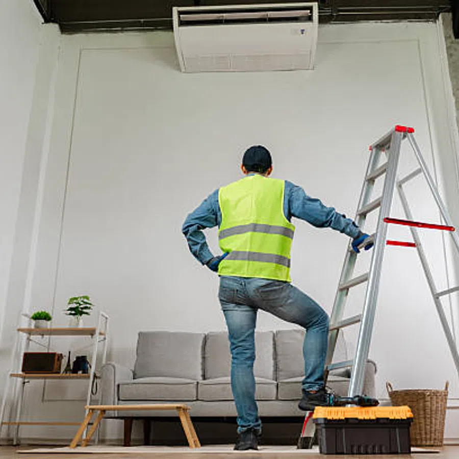 Technician in safety vest standing near ladder and tools, inspecting wall-mounted air conditioner in a modern living room.