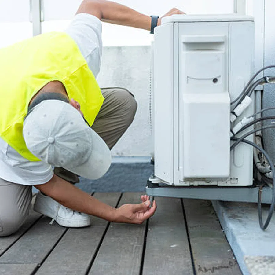 Technician in yellow vest inspecting and repairing an outdoor air conditioning unit on a rooftop deck.