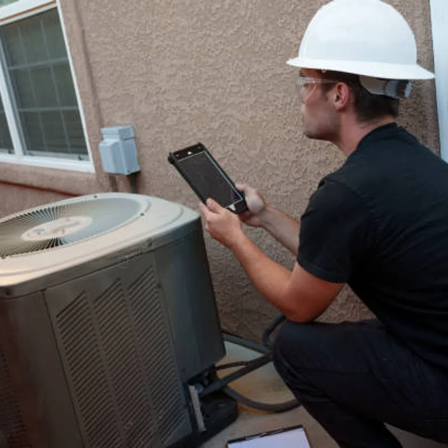 Technician in hard hat inspecting outdoor air conditioning unit using tablet device outside house